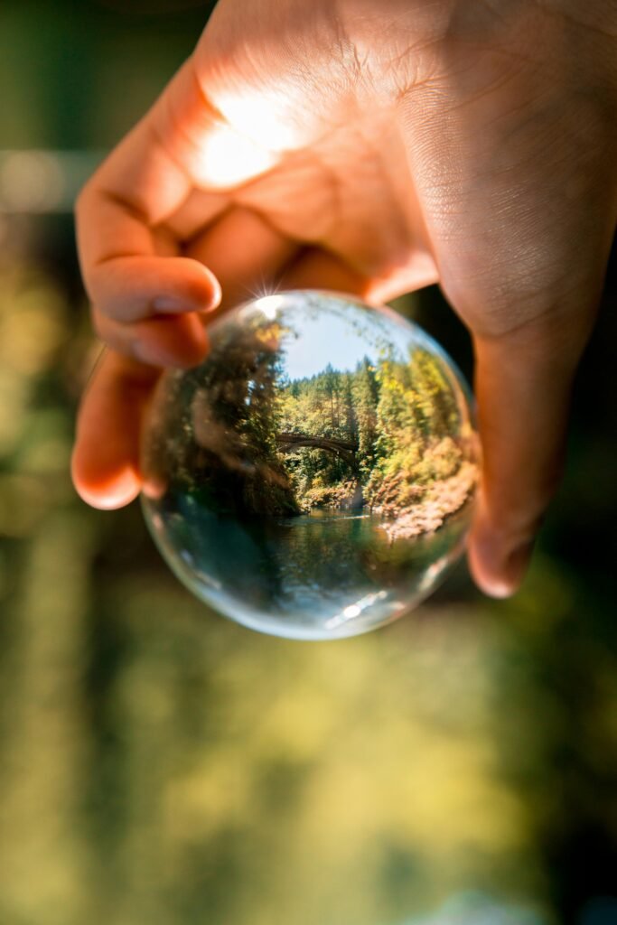 A hand holds a crystal ball reflecting a forest scene with a bridge, creating a unique perspective.